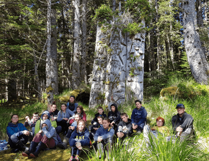 Personnes assises sur l'herbe devant des poteaux totémiques.