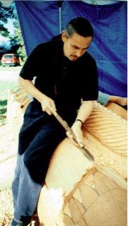 Homme sculptant un totem avec un couteau courbe.