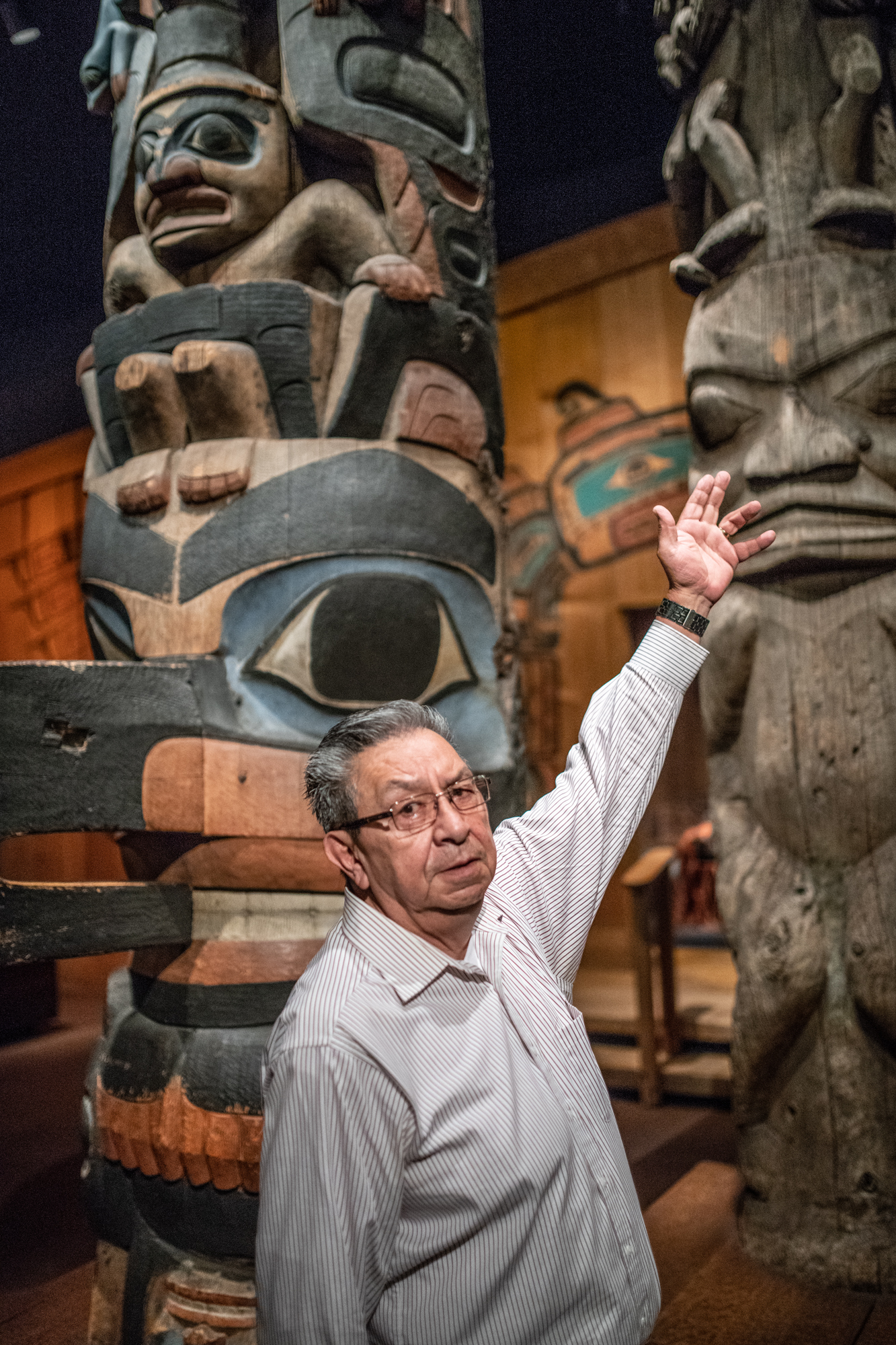 Man stands in front of totem poles, gesturing towards them with his left hand. 