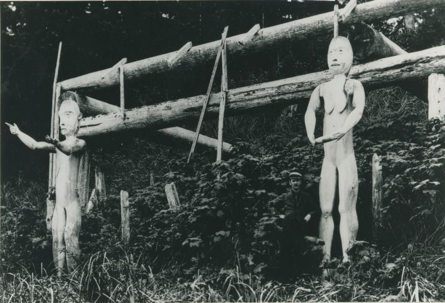 Deux figures humaines sculptées devant une structure de maison.