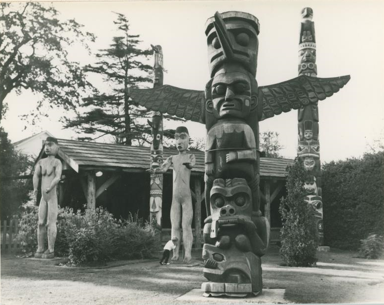 House post standing in Thunderbird Park with the carved human figures in the background with other poles.