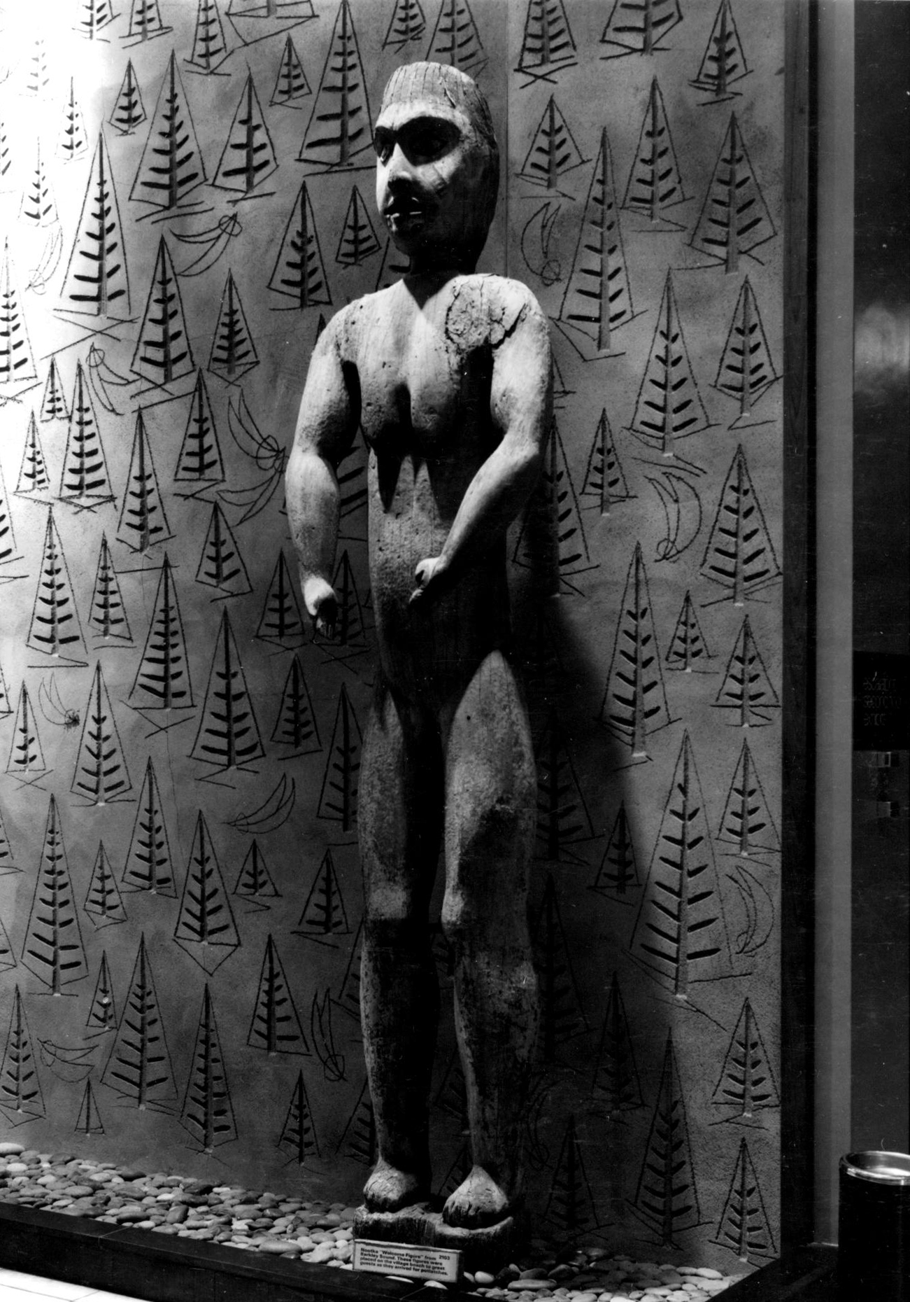 Carved human figure standing inside of the museum against the museum lobby’s wall.