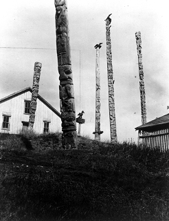 Five poles and a carved figure standing on a hillside with a house in the background.