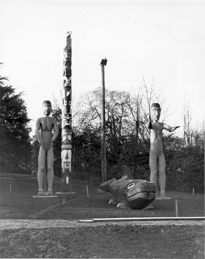 Carved human figures standing in Thunderbird Park with other carved figures and poles.