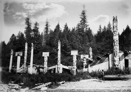 Three houses in Cumshewa village with multiple poles standing in front of them.