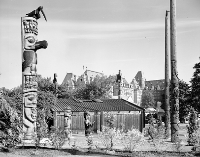 Side view of the pole standing in Thunderbird Park with other poles, carved figures, wooden structure, and The Empress Hotel in the background. 