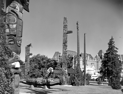 Replica memorial pole standing in Thunderbird Park surrounded by other poles and carvings.