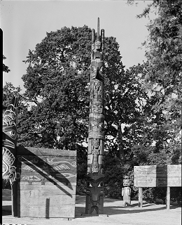 Memorial pole standing in Thunderbird Park between painted screen board and mortuary figure. 