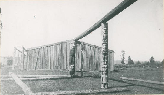House frame with carved house posts, with standing house in background.